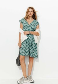 Robe verte à motifs avec un décolleté en V, des manches courtes et des détails de boutons. Associée à des baskets argentées et à une légère veste drapée sur l'épaule.