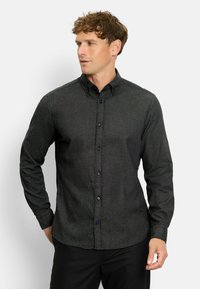 Mann mit lockigem Haar, der ein dunkelgraues, langärmliges Hemd mit Knöpfen und schwarze Hosen trägt. Er schaut nach links, seine Hände sind teilweise sichtbar.