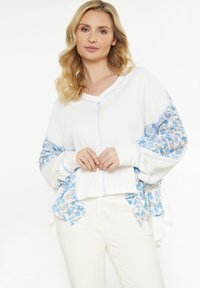 Donna bionda con pantaloni bianchi e un maglione bianco largo, dotato di inserti in tessuto a motivi floreali blu sulle maniche e sui lati.