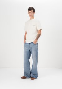 Lys beige bomull t-skjorte med brystlomme, kombinert med løstsittende lyseblå denim jeans og brune sko. Enkel design, avslappet passform.