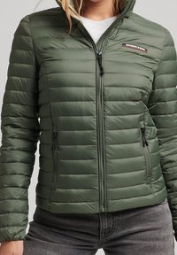 Person som bär en grön vadderad zip-up jacka med två dragkedjefickor och en liten rektangulär logotyp på bröstet.
