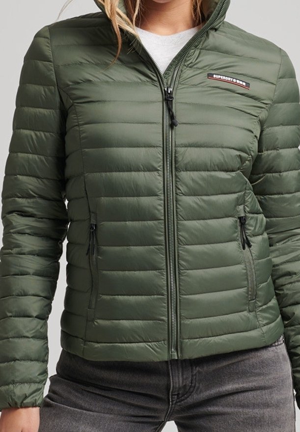 Person som bär en grön vadderad zip-up jacka med två dragkedjefickor och en liten rektangulär logotyp på bröstet.