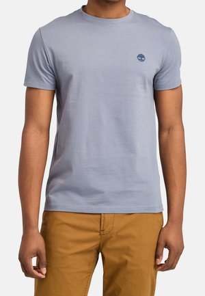 Muž nosící světle šedé tričko s kulatým výstřihem a krátkými rukávy, s malým tmavým logem na levé straně hrudi a hořčicově zbarvené kalhoty.