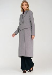 Szary płaszcz w wzór hounds-tooth o dopasowanym kroju, podwójnie zapinany, z wyłogami i dwoma bocznymi kieszeniami. Nosi się go na ciemne rozszerzane spodnie.