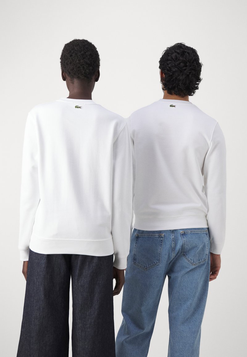 Sudaderas blancas de cuello redondo llevadas por dos personas. Una lleva pantalones anchos de mezclilla oscura; la otra tiene jeans de un azul claro. Pequeño logotipo en el cuello.