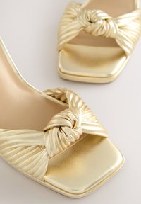 Zlaté sandále na vysokom podpätku so zapleteným dizajnom, s plisovanou textúrou a štvorcovou špičkou. Hladký kovový povrch zdôrazňuje vzhľad.