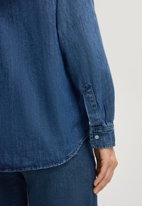 Camicia di denim blu scuro con maniche lunghe. Caratterizzata da un polsino abbottonato e un orlo leggermente curvato. Tessuto morbido e di peso medio.