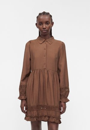 Frau trägt ein braunes Kleid mit langen Ärmeln und Knöpfen vorne, Spitzen-Details an Ärmeln und Saum und Rüschen am unteren Saum.