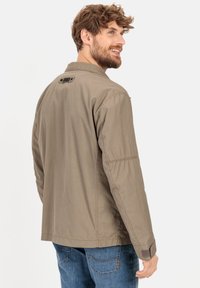 Beige, leichte Jacke mit Kragen, Knopfdetails und gesteppten Akzenten. Getragen mit blauen Jeans, von hinten gezeigt.