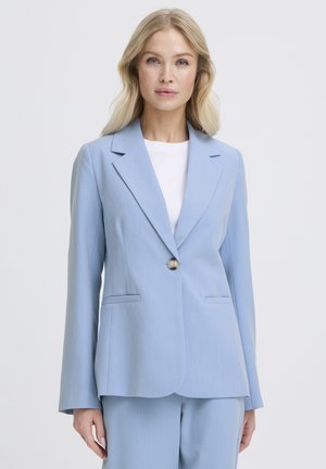 Donna con capelli biondi che indossa un blazer azzurro chiaro con un bottone sopra una maglietta bianca, in piedi davanti a uno sfondo bianco uniforme.