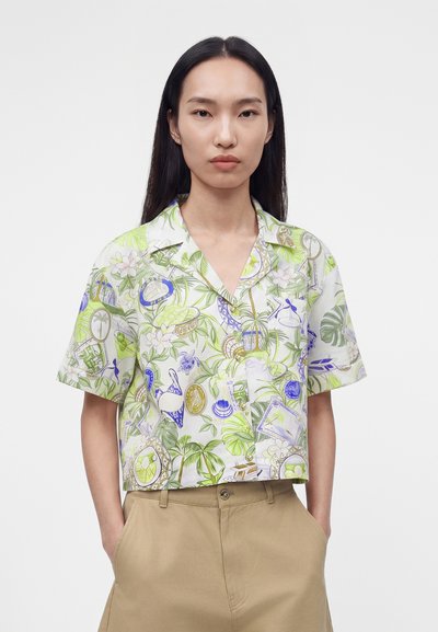 Νέα γυναίκα με μακριά σκούρα μαλλιά φοράει ένα πουκάμισο με κοντό μανίκι και κουμπιά με πράσινο και μοβ τροπικό σχέδιο και μπεζ παντελόνι, με τα χέρια στις τσέπες.