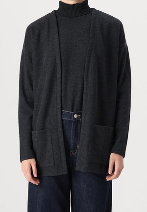 Osoba ubrana w ciemnoszary rozpinany kardigan na pasującym golfie oraz ciemnoniebieskie dżinsy, z rękami swobodnie opuszczonymi wzdłuż ciała.