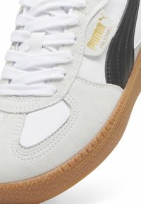 Baskets Puma Palermo blanches et grises avec une bande noire, logo Puma doré, lacets blancs et semelle en caoutchouc gomme, gros plan sur l'avant et le côté.