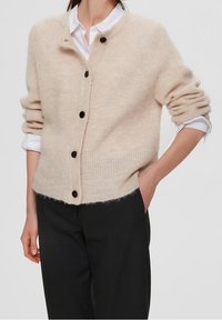 Cardigan lavorato a maglia beige con finitura testurizzata, scollatura rotonda e bottoni neri, abbinato a una camicia bianca e pantaloni neri.