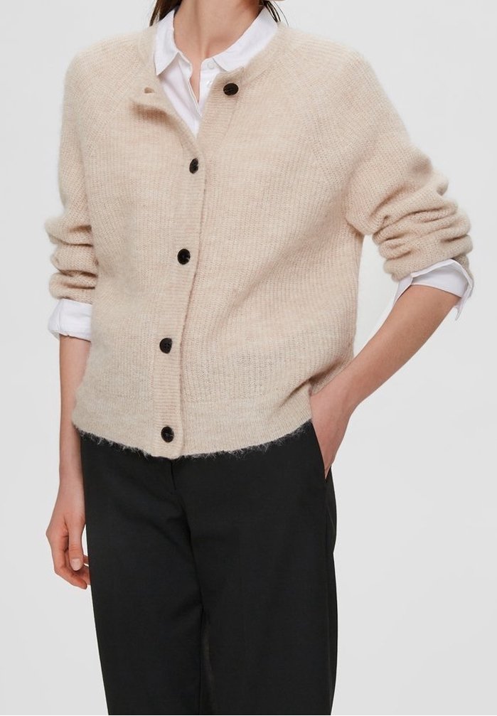 Cardigan lavorato a maglia beige con finitura testurizzata, scollatura rotonda e bottoni neri, abbinato a una camicia bianca e pantaloni neri.