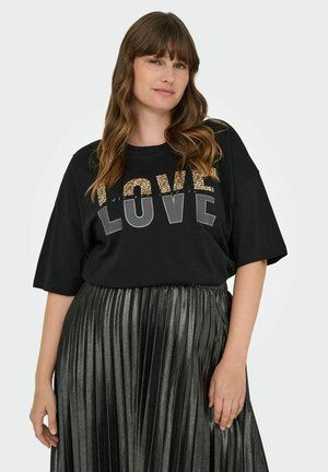 Vrouw met golvend bruin haar, gekleed in een oversized zwart T-shirt met de tekst "LOVE" en een metalen geplooide rok, staande tegen een effen achtergrond.