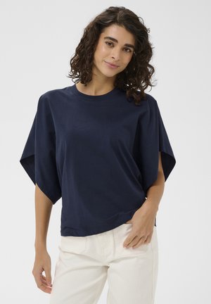 Femme aux cheveux bouclés portant un haut bleu marine à manches larges et un pantalon blanc, posant avec une main dans la poche devant un fond uni.