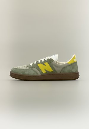Low-Top New Balance Sneaker aus grünem Wildleder und Beige mit gelben Logoakzenten und brauner Sohle, seitlich auf neutralem Hintergrund abgebildet.