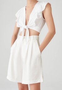 Mujer lleva pantalones cortos acampanados de talle alto blanco y un top corto sin mangas blanco con volantes en los hombros atado en el frente.