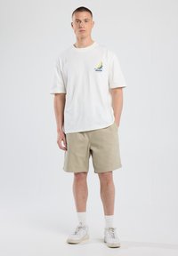 Camiseta blanca de algodón con un gráfico azul y amarillo de "Atlanta" en el pecho, combinada con pantalones cortos beige y zapatos deportivos blancos.