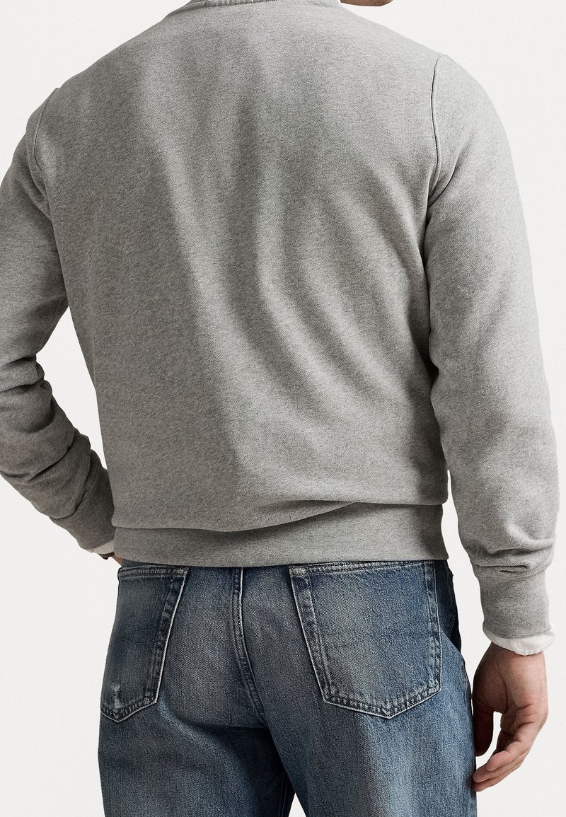 Homme portant un sweat-shirt gris clair et un jean en denim bleu, vu de dos, main reposant sur la hanche sur un fond uni.