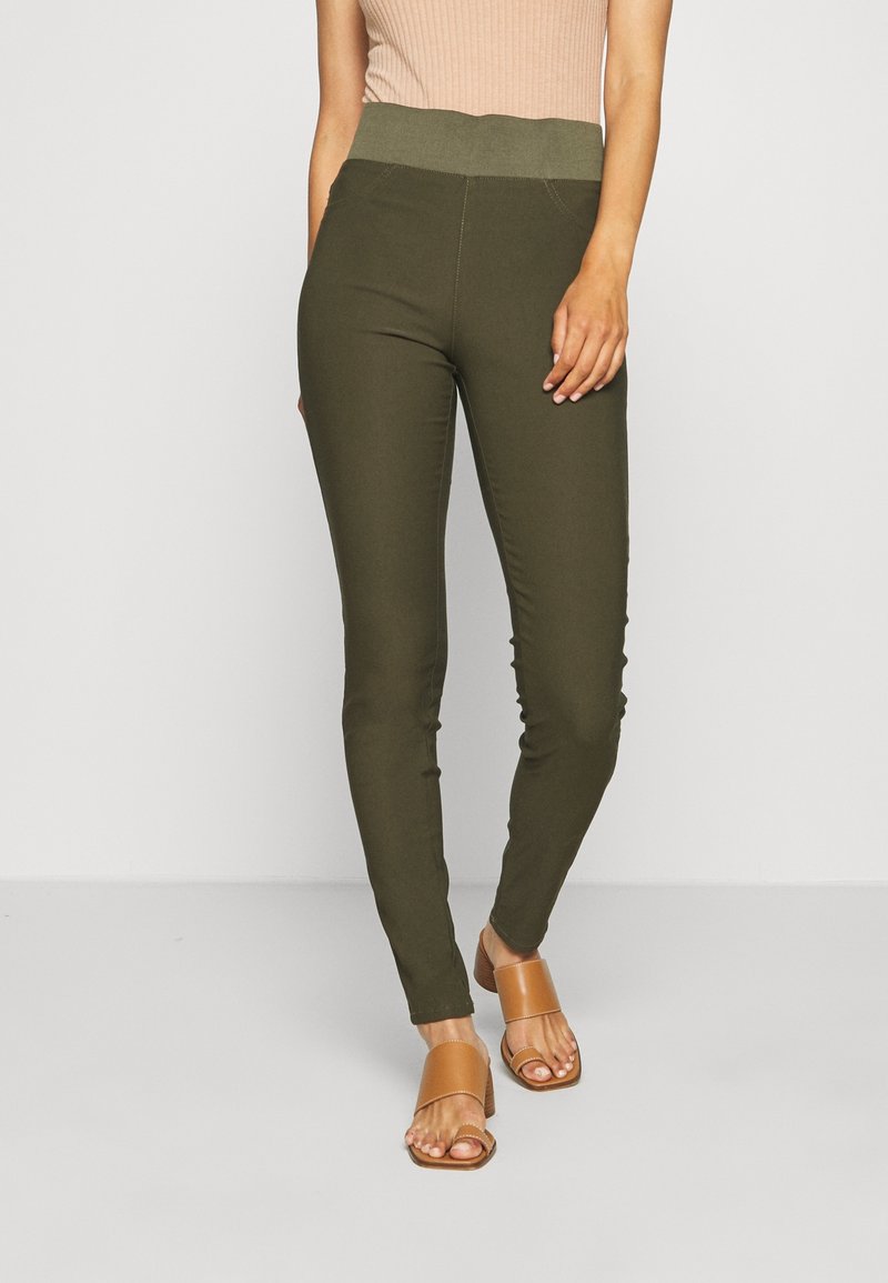 Femme portant un pantalon slim vert olive avec une large ceinture élastique et des sandales à talons beige, debout sur un fond clair uni.