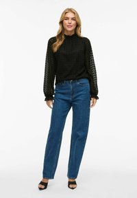 Maglione nero a maniche lunghe con scollo arricciato, abbinato a jeans dritti in denim blu a vita alta e sandali neri con punta aperta.