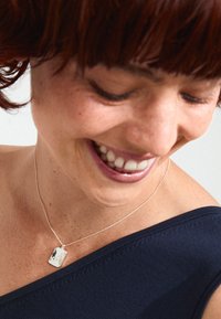 Silverhalsband med ettrektangulär form, med en slät yta och subtil lyster, bärs på en silverkedja mot en marinblå topp.