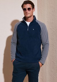 Navy blue fleece pullover with gray raglan sleeves, front zipper, and a small logo on the chest. Soft texture with a relaxed fit.