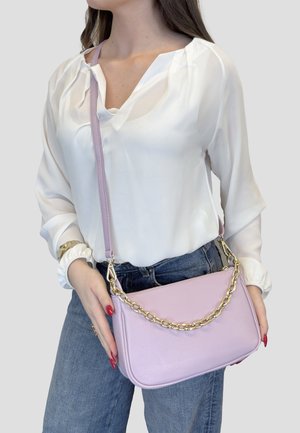 Frau in weißer Bluse und blauen Jeans hält eine hellviolette Handtasche mit goldener Kette und Schulterriemen.