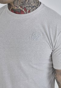Svijetlosivi kratki rukav majica od mekanog materijala, s diskretnim izvezenim logotipom na lijevoj strani grudnog koša. Glatka tekstura, klasičan kroj.