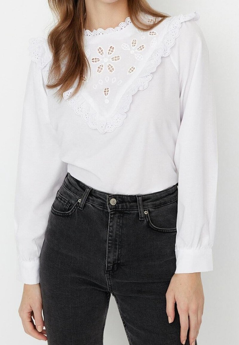 Femme portant un chemisier blanc à manches longues avec un détail en dentelle œillets florale et un col à volants, associé à un jean noir taille haute.