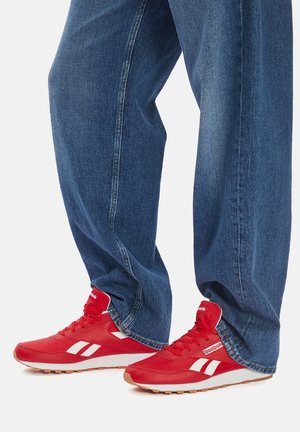 Zapatillas deportivas rojas con detalles y logotipo blancos, con textura suave, punta redonda y suela acolchada, combinadas con pantalones vaqueros azules holgados.