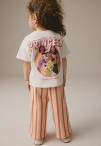Camiseta blanca con un colorido gráfico de princesa en la espalda, combinada con pantalones sueltos de rayas naranjas. Tejido de algodón suave, corte relajado.