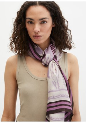 Młoda kobieta o kręconych włosach, ubrana w beżowy top na ramiączkach i fioletowy wzorzysty szalik zawiązany wokół szyi, na jednolitym tle.