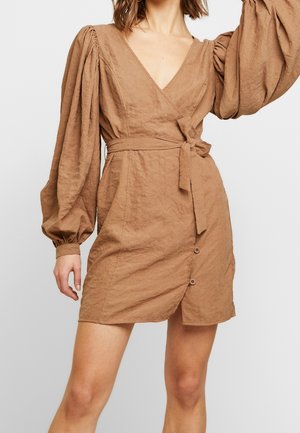Femme portant une robe portefeuille marron clair avec de longues manches bouffantes, un col en V, des détails de boutons et une ceinture nouée à la taille sur un fond uni.
