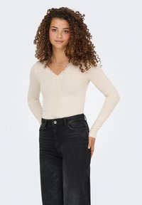 Kerman ribbattu pitkähihaine paita V-pääntie ja pitsireunus pääntien ympäri, yhdistettynä korkeavyötäröisiin mustiin farkkuihin.