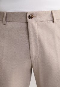 Beige byxor med en strukturerad väv, som har knappstängning och sidofickor. Tyget verkar lätt och hållbart.