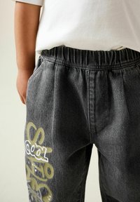 Graue Denim-Hose mit lockerer Passform, ausgestattet mit einem elastischen Bund und Seitentaschen. Ein auffälliger Kunststoff-Print in Gelb und Weiß verziert das Bein.