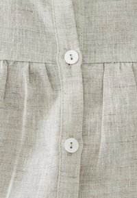 Světlá šedá lněná tkanina s texturovanou vazbou, která má dva bílé knoflíky a detail s nasbíranou plisováním pod légu knoflíků.