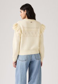 Pull en maille jaune clair avec des accents de volants sur les épaules, motif en dentelle complexe dans le dos, ourlet et poignets côtelés, associé à un jean en denim.