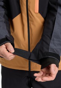 Person, der den Ärmel einer schwarz-grauen Jacke mit einem Klettverschluss anpasst, während er eine braun-schwarze Zip-Jacke trägt.