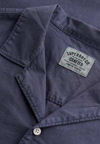 Col de chemise bleu foncé avec un bouton blanc et une étiquette en tissu blanc portant l'inscription "Superdry Co. Crafted" cousue à l'intérieur près de l'encolure.