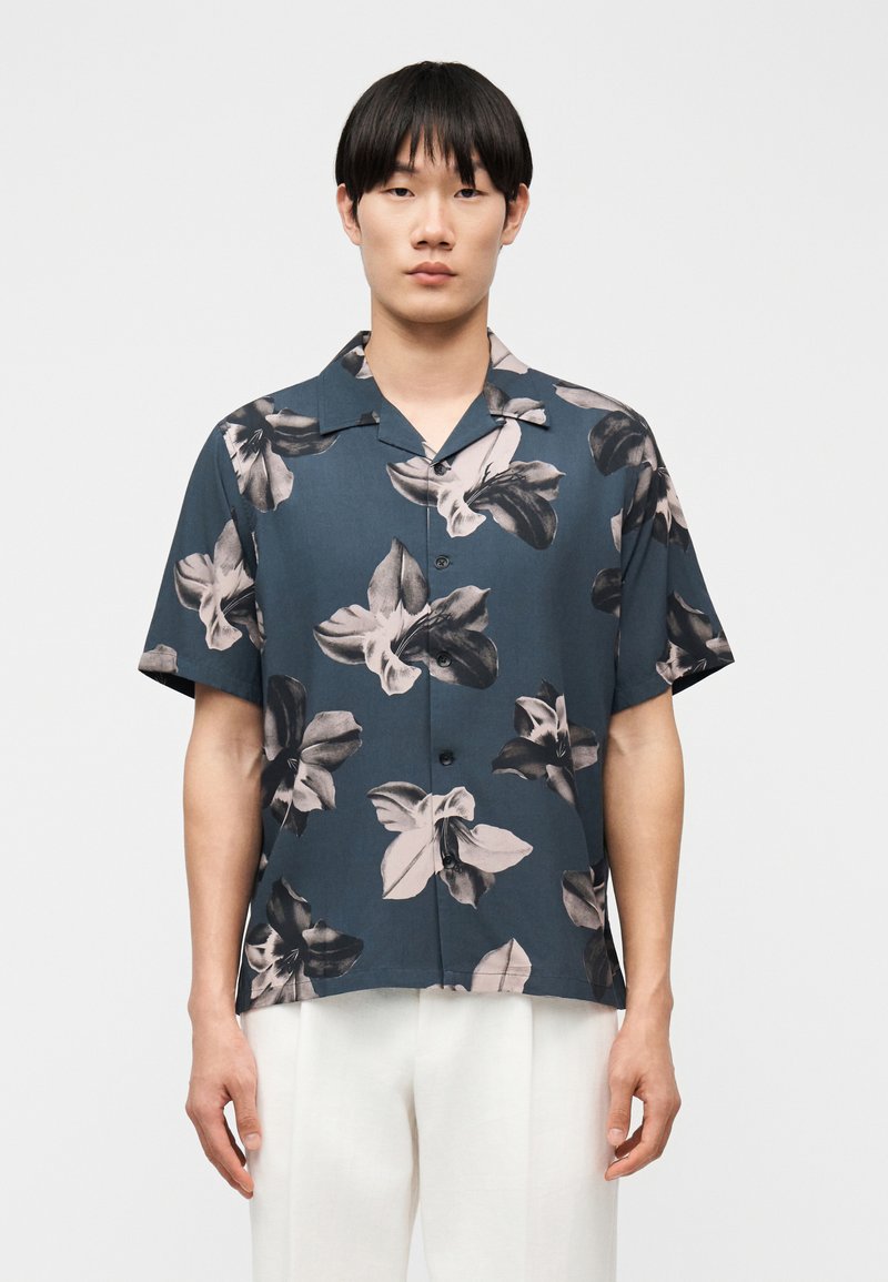 Jeune homme portant une chemise à manches courtes bleu marine avec un grand imprimé floral noir et blanc, et un pantalon blanc, debout devant un fond uni.