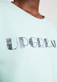 T-shirt azul claro feito de tecido suave, apresentando um grande gráfico texturizado em cores escuras com a palavra "UPGREAT". Gola redonda com mangas curtas.