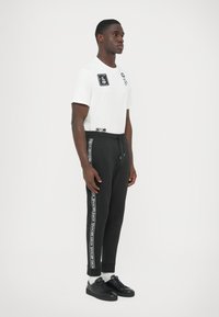 Biała koszulka z czarnymi graficznymi detalami w połączeniu z czarnymi spodniami dresowymi z bocznymi taśmami z logo. Model nosi czarne trampki i szare skarpetki.