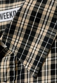 Chemise à carreaux mettant en avant des couleurs noir et beige avec des accents blancs. Tissu en coton, manches courtes et fermeture à boutons sont visibles.