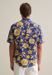Camisa de manga corta en azul marino con un patrón floral amarillo; tela suave; corte relajado; dobladillo curvado; collar minimalista y detalles de botones.