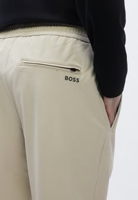 Bézs színű nadrág sima anyagból, elasztikus derékrésszel, cipzáras hátsó zsebbel és fekete "BOSS" logóval nyomtatva.