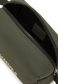 Olivově zelená crossbody taška z hladkého materiálu, se zipovým uzávěrem a značkovou etiketou. Má texturovaný vnitřek a kontrastní šití.